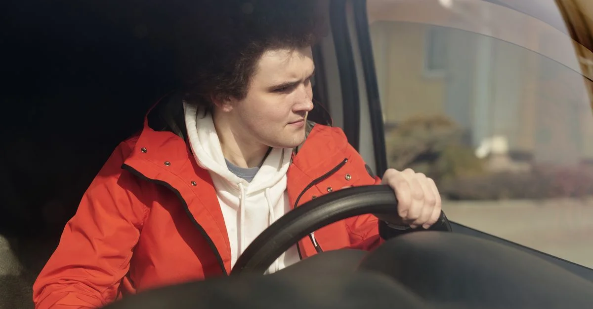 Un jeune homme en veste rouge dirige une voiture avec concentration, profitant d'une conduite paisible.