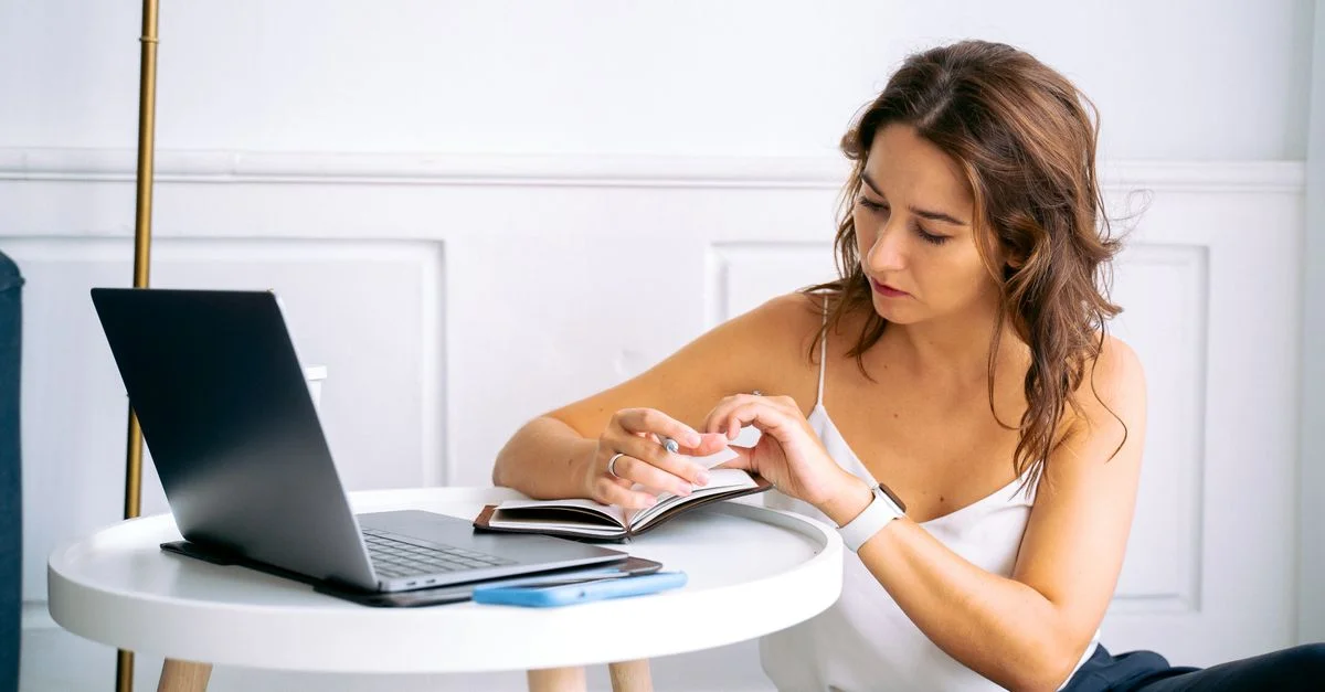 Une femme en haut blanc écrit dans un carnet à une petite table avec un ordinateur portable.