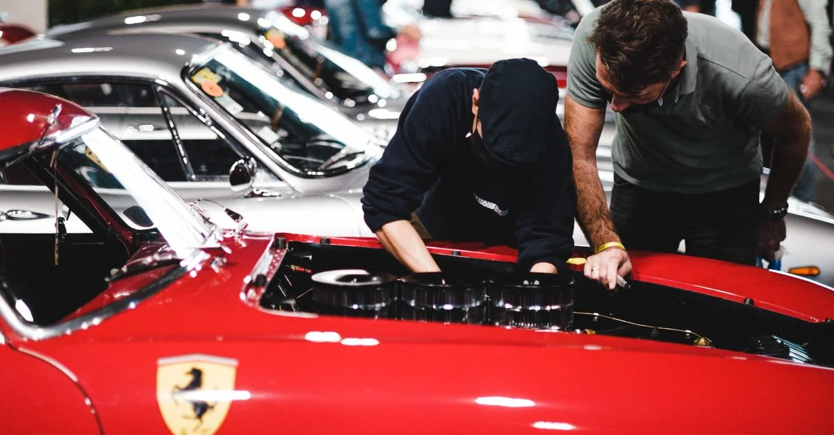 Ferrari rouge avec des mécaniciens inspectant le moteur lors d'un salon de voitures classiques.