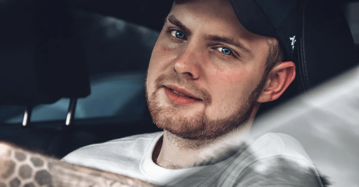 Gros plan d'un homme souriant, barbu et aux yeux bleus assis dans une voiture, créant une ambiance détendue.