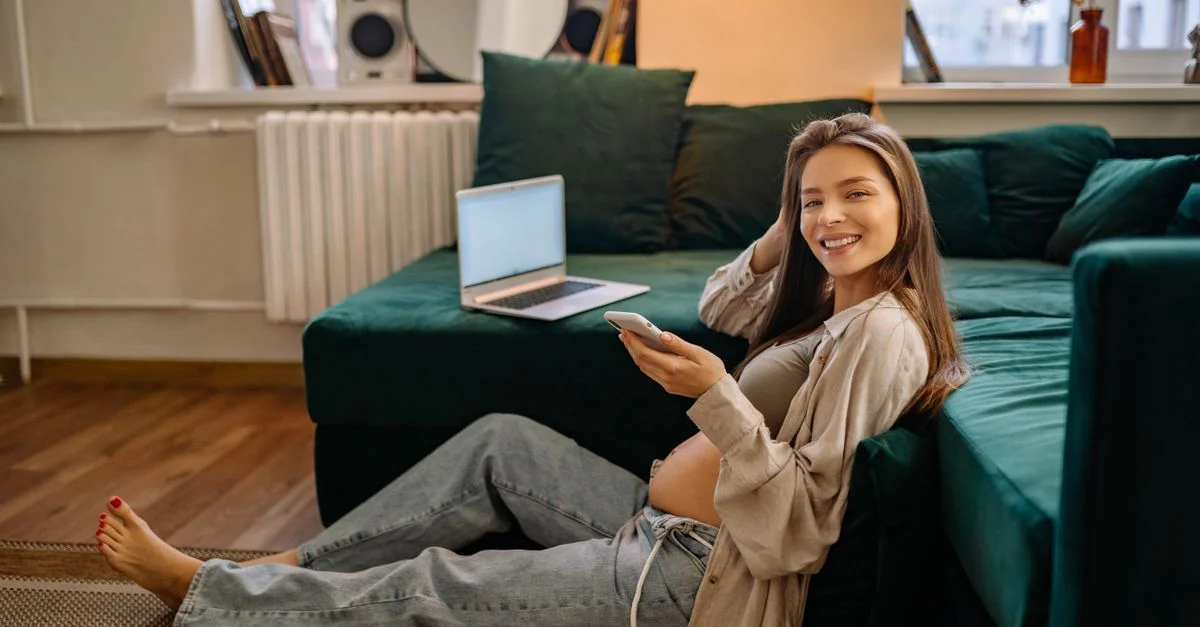 Femme enceinte souriante assise par terre utilisant un smartphone à l'intérieur.