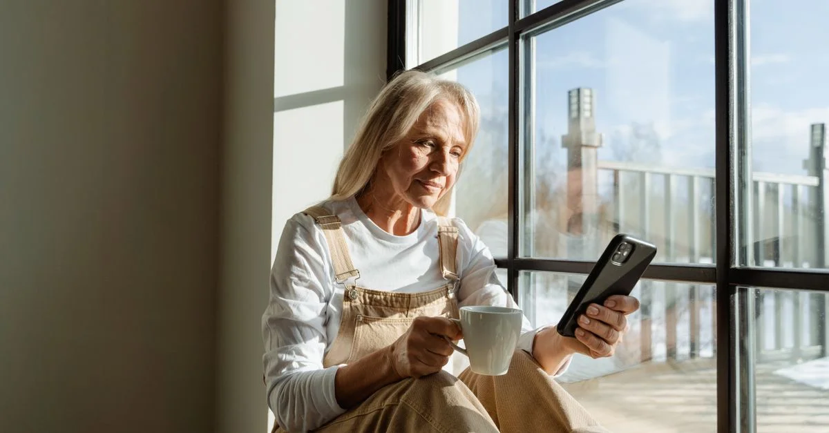 Femme âgée se détendant avec un café et un smartphone près d'une fenêtre ensoleillée.