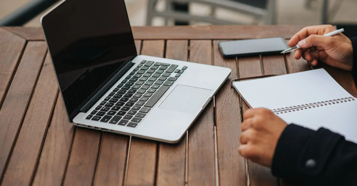 Ordinateur portable ouvert et une personne écrivant dans un carnet sur une table en bois, prête pour le travail à distance.