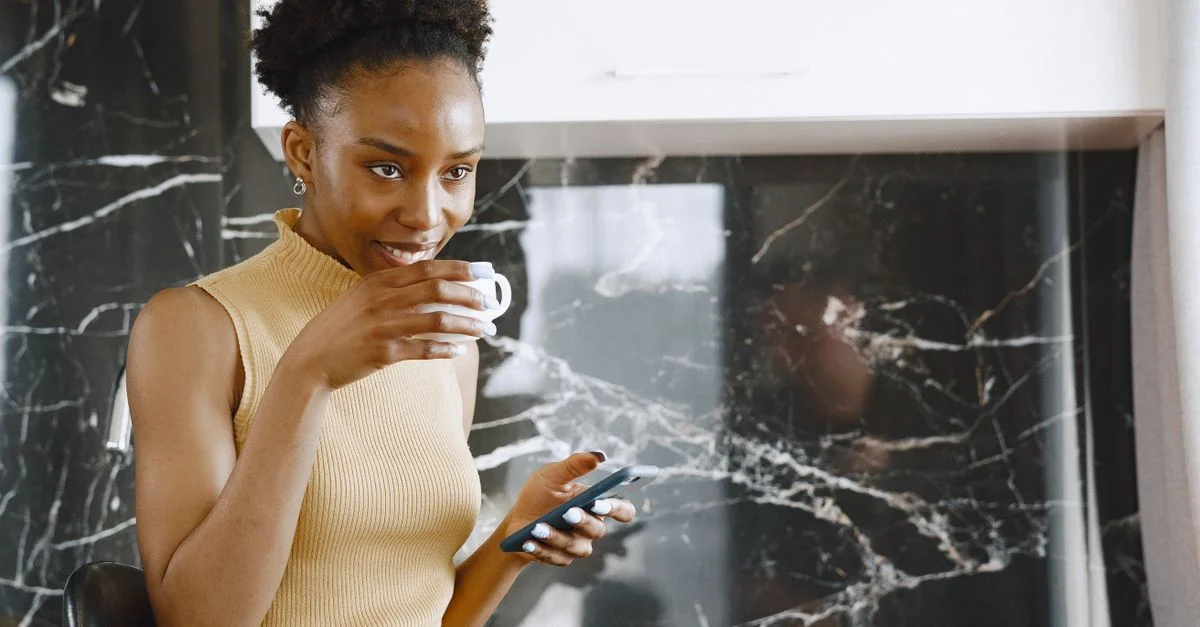 Femme africaine avec des cheveux afros, buvant du café et utilisant un smartphone à l'intérieur, souriante et détendue.